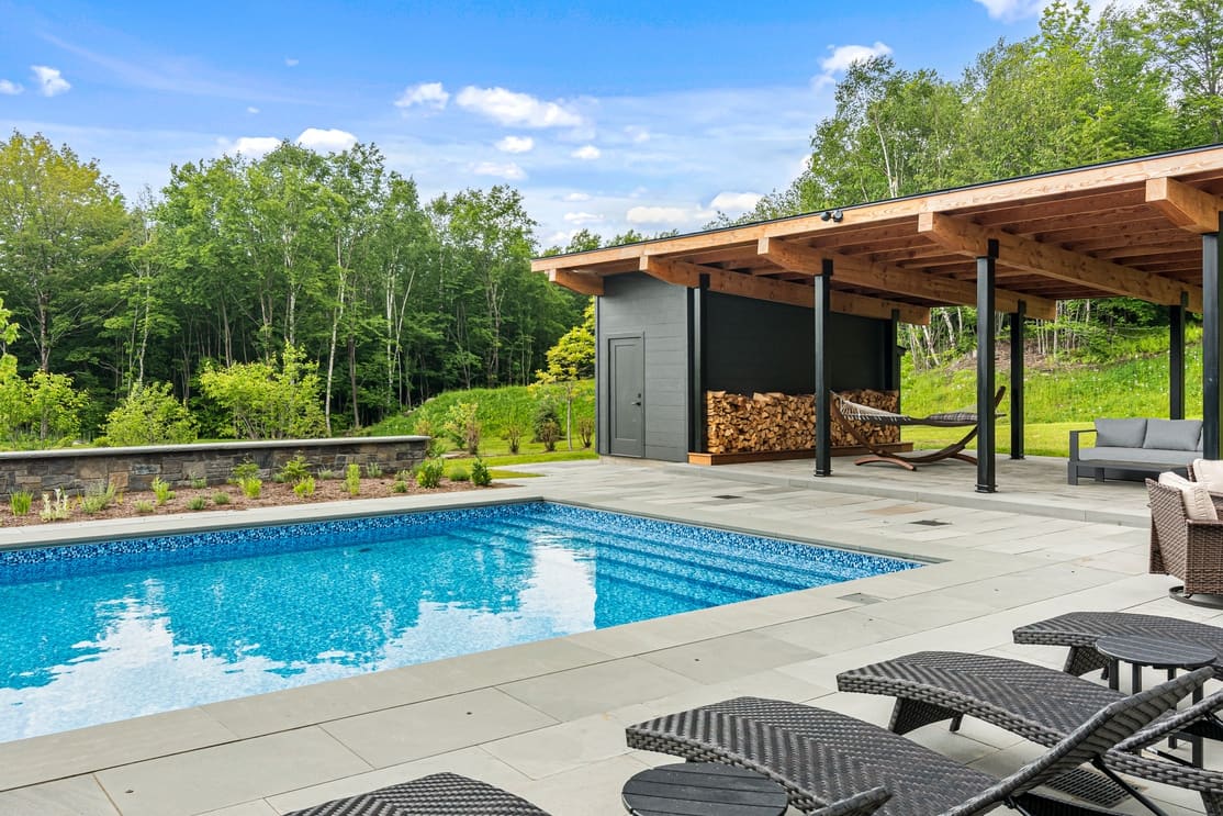 Modern backyard pool with stone patio, covered pavilion, seating, and stacked firewood, showcasing landscape design-build and outdoor craftsmanship.