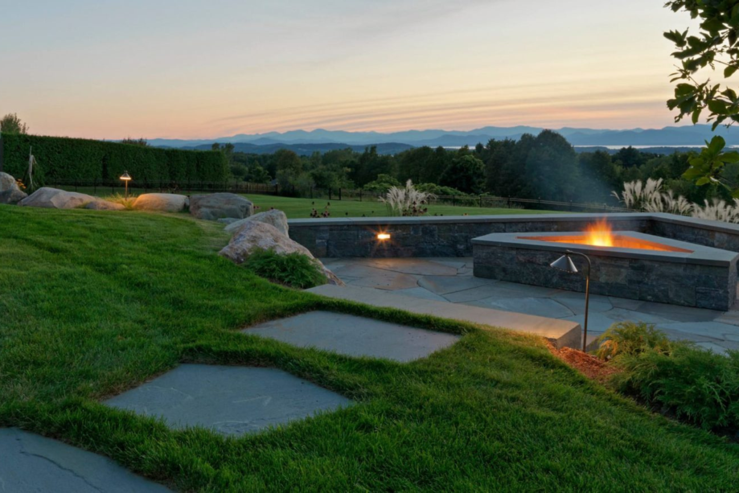 Outdoor patio with a fire feature, stone walkway, and a landscaped lawn at sunset.