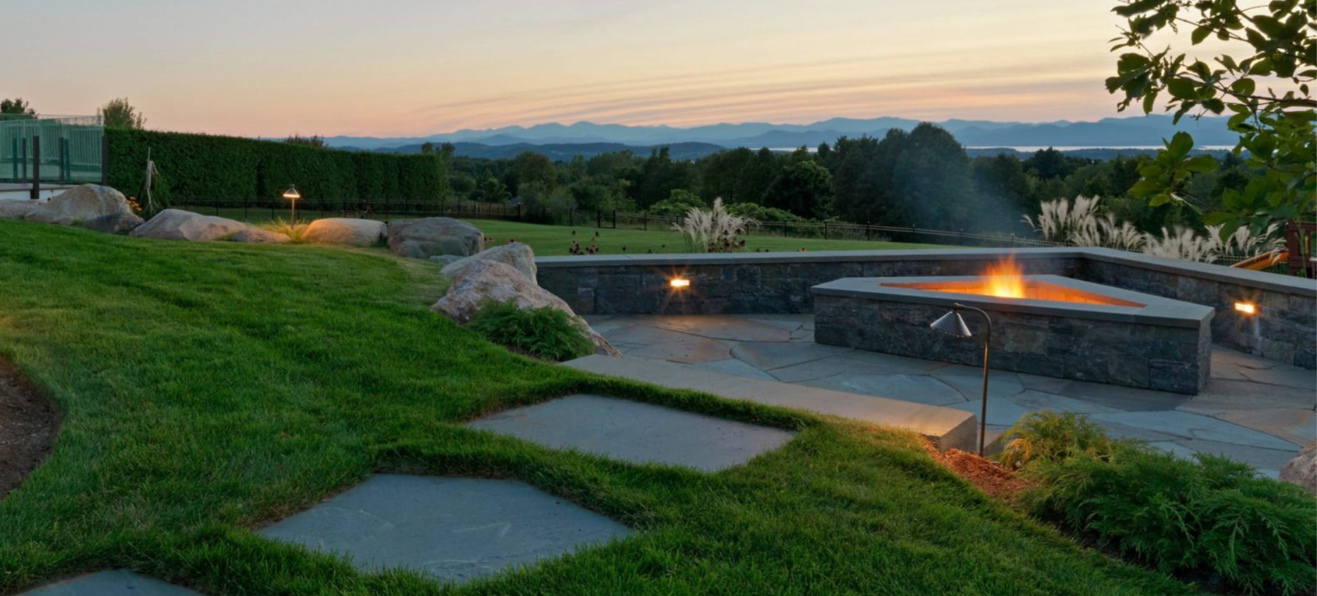 Outdoor patio with a fire feature, stone walkway, and a landscaped lawn at sunset.